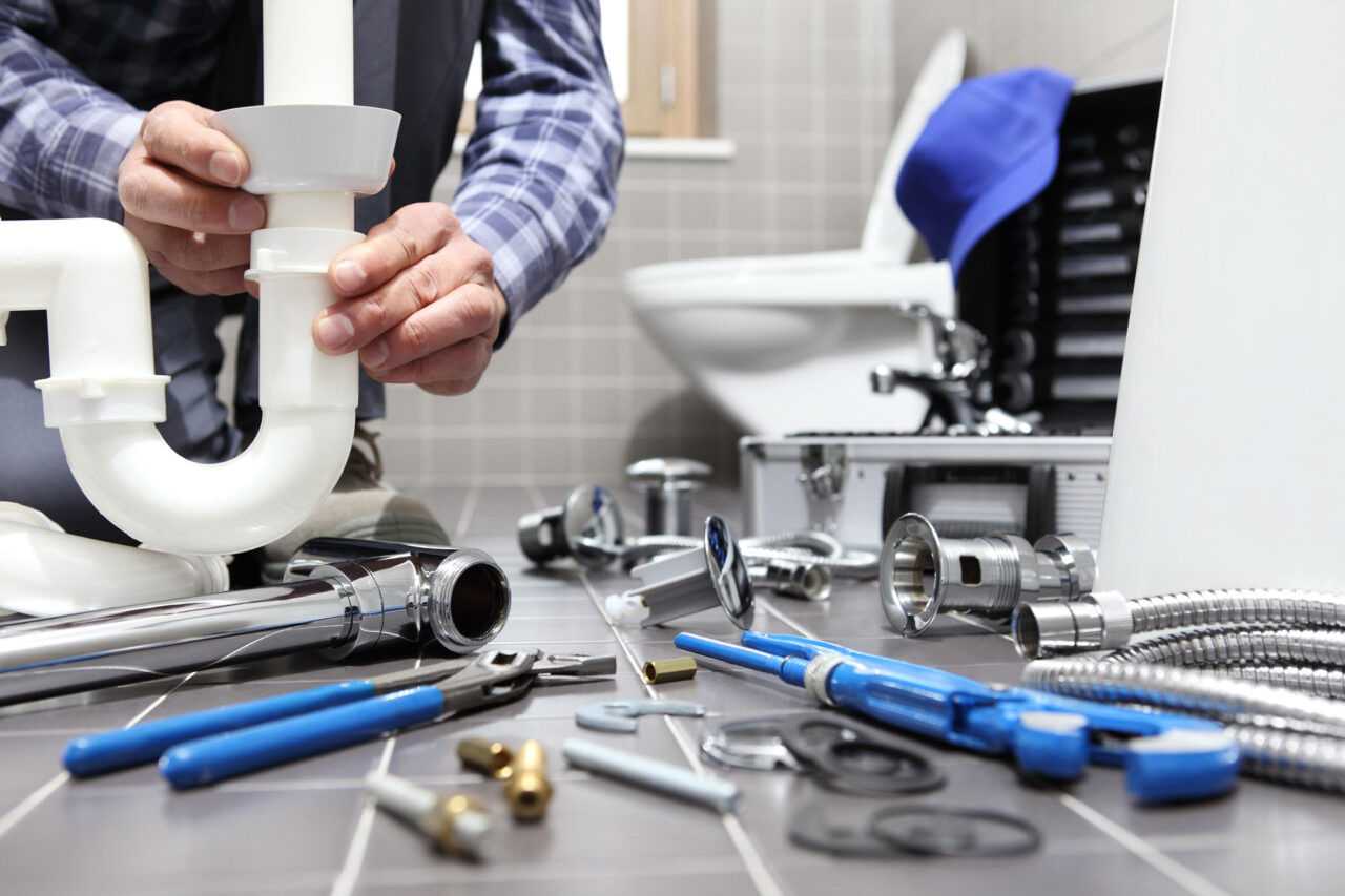 Plumber fixing sink pipes in a bathroom, surrounded by tools and plumbing parts.