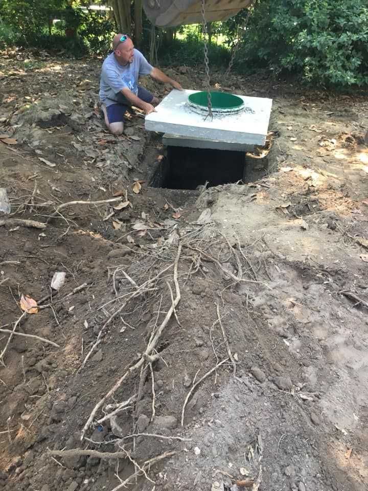 Man installing a septic tank lid in a backyard, surrounded by dirt and roots.